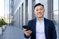 Close-up portrait of a smiling and successful Asian businessman standing near an office building, holding a mobile phone Royalty Free Stock Photo