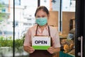 Close up portrait smiling young Asian woman in a beige shirt with a apron wearing a face mask holding an open sign board in front Royalty Free Stock Photo