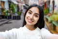 Close-up portrait of a smiling young Indian woman in a white sweater taking a selfie on a city street, talking online to Royalty Free Stock Photo