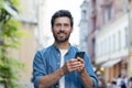 Close-up portrait of a smiling young man in a blue shirt standing on a city street, holding a mobile phone and looking Royalty Free Stock Photo