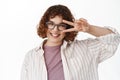 Close up portrait of smiling young natural girl with curly hair, wearing glasses, showing peace kawaii v-sign near eye