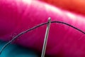 A close up portrait of some sewing thread put through the hole of a needle in front of an out of focus spool or roll of yarn. The