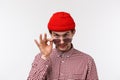 Close-up portrait of suspicious funny young guy in red beanie and checked shirt, take-off sunglasses and look from under Royalty Free Stock Photo