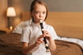 Close-up portrait of talented adorable little girl playing flute sitting on bed in bedroom, looking at camera. Royalty Free Stock Photo