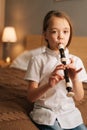 Close-up portrait of talented adorable little girl playing flute sitting on bed in bedroom, looking at camera. Royalty Free Stock Photo