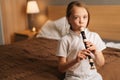 Close-up portrait of talented adorable little girl playing flute sitting on bed in bedroom, looking at camera. Royalty Free Stock Photo