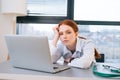 Close-up portrait of tired exhausted young female doctor in white coat sitting at desk with laptop and looking at camera Royalty Free Stock Photo