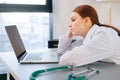 Close-up portrait of tired exhausted young female doctor in white coat sitting at desk with laptop and looking at screen Royalty Free Stock Photo