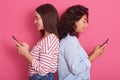 Close up portrait of two cute young women pose together  over pink background while standing back to back to each other, Royalty Free Stock Photo