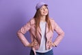 Close up portrait of urban portrait of stylish sportive girl posing against lilac wall, wearing hat, shirt, jacket, holding hands