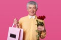 Close up portrait of white haired elderly man in yellow shirt and white bow tie, holds red roses and bags with present, being in