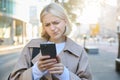 Close up portrait of woman looking sceptical at her mobile phone, standing on street, frowning and standing perplexed