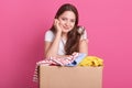 Close up portrait of woman separates clothes for donation box, standing isolated over pink studio background, attractive female