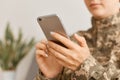 Close up portrait of woman soldier wearing camouflage uniform sitting with smart phone in hands, using cell phone, checking social