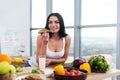 Close-up portrait of woman standing in kitchen, leaning on wooden table, having snack. Smiling girl maintains healthy Royalty Free Stock Photo