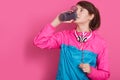 Close up portrait of woman wearin blue and rose sportswear drinking water from bottle, model posing over rosy background
