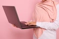 Close-up portrait of a woman wearing pink hijab standing against colored background with an open laptop in hands and typing text. Royalty Free Stock Photo