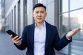 Close-up portrait of a young Asian male businessman standing near an office building and upset, spreading his hands Royalty Free Stock Photo