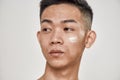 Close up portrait of young asian man with problematic skin applied cream on his face, looking aside isolated over white