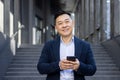 Close-up portrait of a young Asian man in a suit standing outside an office building, holding a phone and smiling at the Royalty Free Stock Photo