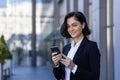 Close-up portrait of a young business woman standing near an office building, holding a phone and smiling at the camera Royalty Free Stock Photo