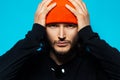 Close-up portrait of young confident man, holding hands on head. Wearing orange hat and black sweater. Studio photo. Royalty Free Stock Photo