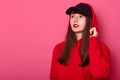 Close up portrait of young cute brunette teenager girl in casual red hoodie and cap, posing with bright listick on pink background Royalty Free Stock Photo