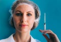 Close-up portrait of young female doctor in medical cap and white gown and syringe on her hand on blue background. Royalty Free Stock Photo