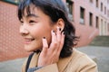 Close up portrait of young korean woman touches earphone, listens music in headphones with pleased smiling face, walks Royalty Free Stock Photo