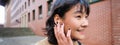 Close up portrait of young korean woman touches earphone, listens music in headphones with pleased smiling face, walks Royalty Free Stock Photo