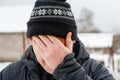 Close-up portrait of young man in warm hat outside on rural winter snowy house background. Tired millennial smiling