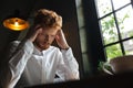 Close-up portrait of young readhead bearded overworked man in white shirt touching his head while sitting at office