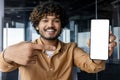 Close-up portrait of young smiling Indian man sitting in office and holding mobile phone camera, pointing finger at Royalty Free Stock Photo