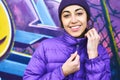 Close-up portrait of young smiling woman wearing in purple down jacket and knitted hat posing against wall painted with Royalty Free Stock Photo