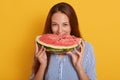 Close up portrait of young woman biting slice of watermelon while looking directly at camera, posing isolated over yellow Royalty Free Stock Photo