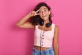 Close up portrait of young woman making victory or peace sign and covering her eye with it, posing over pink wall background Royalty Free Stock Photo