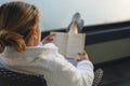 Close up of portrait of young woman in white coat sitting in wicker chair on balcony and reading book Royalty Free Stock Photo