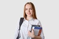 Close up portraitof young girl teenagercarries rucksack, holds colourful folder with papers, has pleasant smile, has lectures in