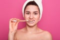 Close up portraitof young smiling woman cleaning her teeth with toothbrush in bathroom, stands wearing white towel and bared