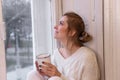 Close-up portraits of young woman sitting on windowsill, drinking cocoa from cup, looking out window