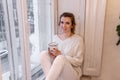 Close-up portraits of young woman sitting on windowsill, drinking cocoa from cup, looking out window