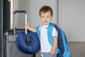 Close-up portrat of cute smiling little baby boy with big backpack and suitcase with travelling pillow stay near open door ready t