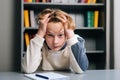 Close-up portriat of exhausted pupil boy tired from studying holding head head with hands while sitting at desk