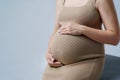 Close-up of a pregnant woman in a beige dress hugging her stomach with both hands on a gray background Royalty Free Stock Photo