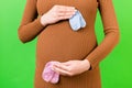 Close up of pregnant woman in brown dress holding baby pink and blue socks against her belly at green background. Is it a boy or a Royalty Free Stock Photo