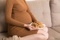 Close up of pregnant woman enjoys eating cookies resting on the sofa at home. Unhealthy sweet pastry during pregnancy concept