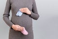 Close up of pregnant woman in gray dress holding baby pink and blue socks against her belly at gray background. Expecting twins. Royalty Free Stock Photo