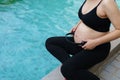 Pregnant woman sitting on edge of swimming pool while applying headphones to her belly for prenatal music stimulation