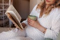 Close up Pregnant woman sitting at home in bed, reading book and drinking tea or water from cup Royalty Free Stock Photo