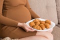 Close up of pregnant woman sitting on the sofa is eating croissants from a plate at home. Unhealthy diet. Control your weight
